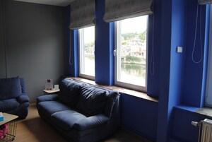 Una habitación con un sofá azul, una mesita y un ventanal con vistas a un edificio y al agua.