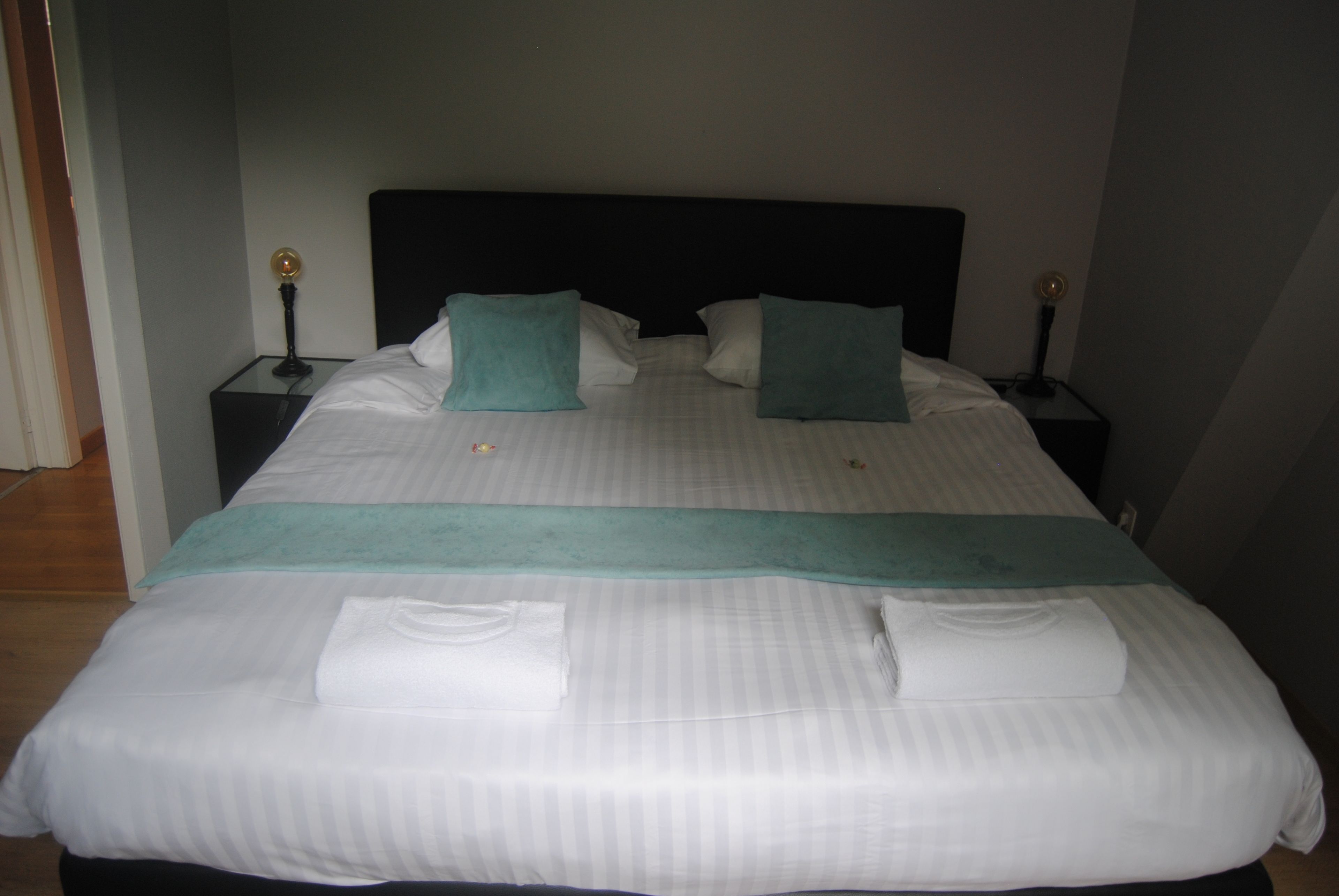 Un lit bien fait, avec du linge de lit blanc, deux coussins turquoise et un plaid turquoise.