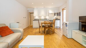 A modern living room with a white sofa, a wooden dining table with chairs, a white coffee table, and a compact kitchen with white cabinets and a built-in microwave. - Cromwell Rooms (Cambridge)