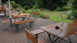 Un espace extérieur aménagé en terrasse, avec des tables et des bancs en bois, entouré de verdure et doté d’un sol en briques.