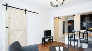 A modern living room with a sliding barn door, a small black TV stand, a round coffee table with two white cups, and a kitchen area with a black brick backsplash and wooden cabinets.
