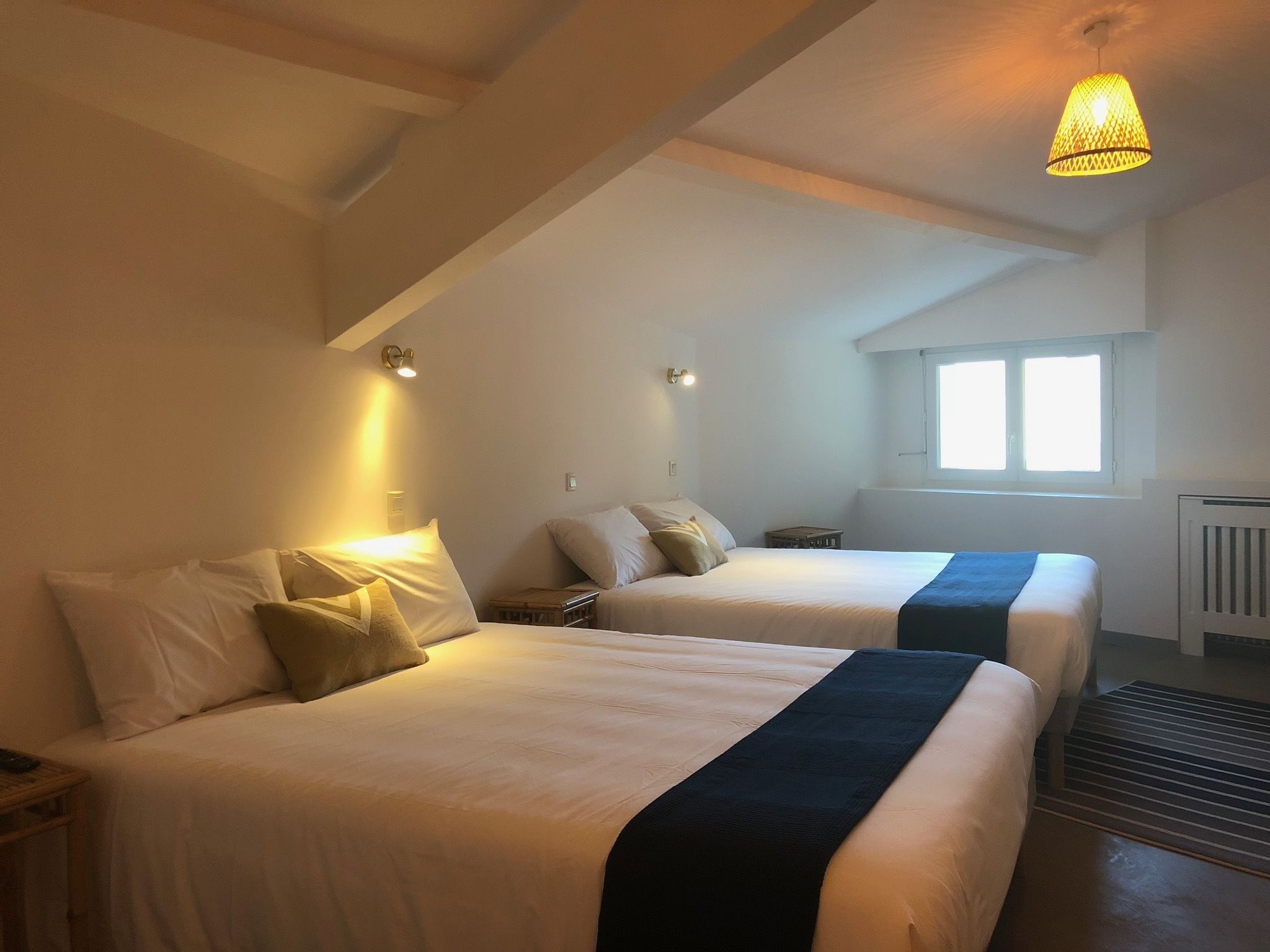 Une chambre avec deux lits, chacun doté de draps blancs et d’une tête de lit sombre. On y trouve des tables de chevet avec des lampes, une fenêtre avec des stores et un radiateur fixé au mur.