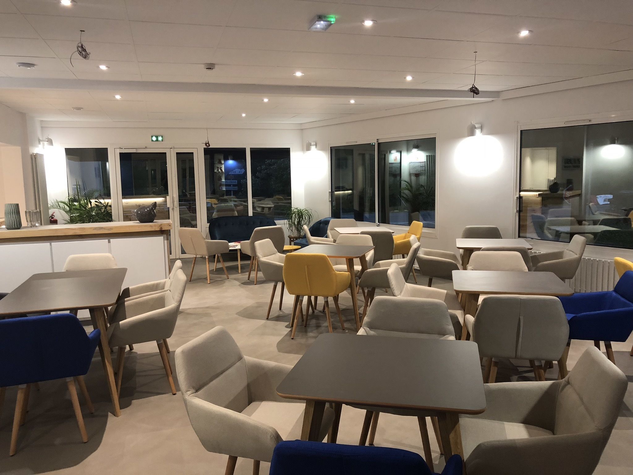 Un espace repas moderne, doté de tables et de chaises, de grandes fenêtres et de plantes d’intérieur.