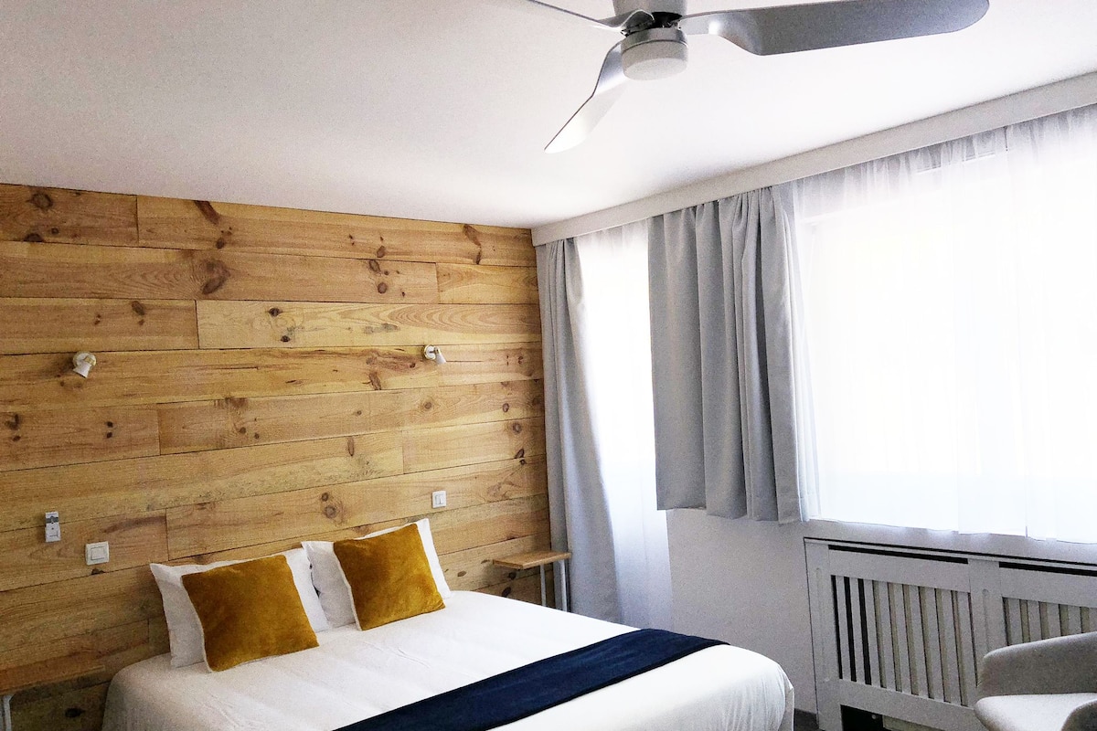 A hotel room with a wooden headboard, a bed with white bedding and gold pillows, a ceiling fan, and a window with curtains.
