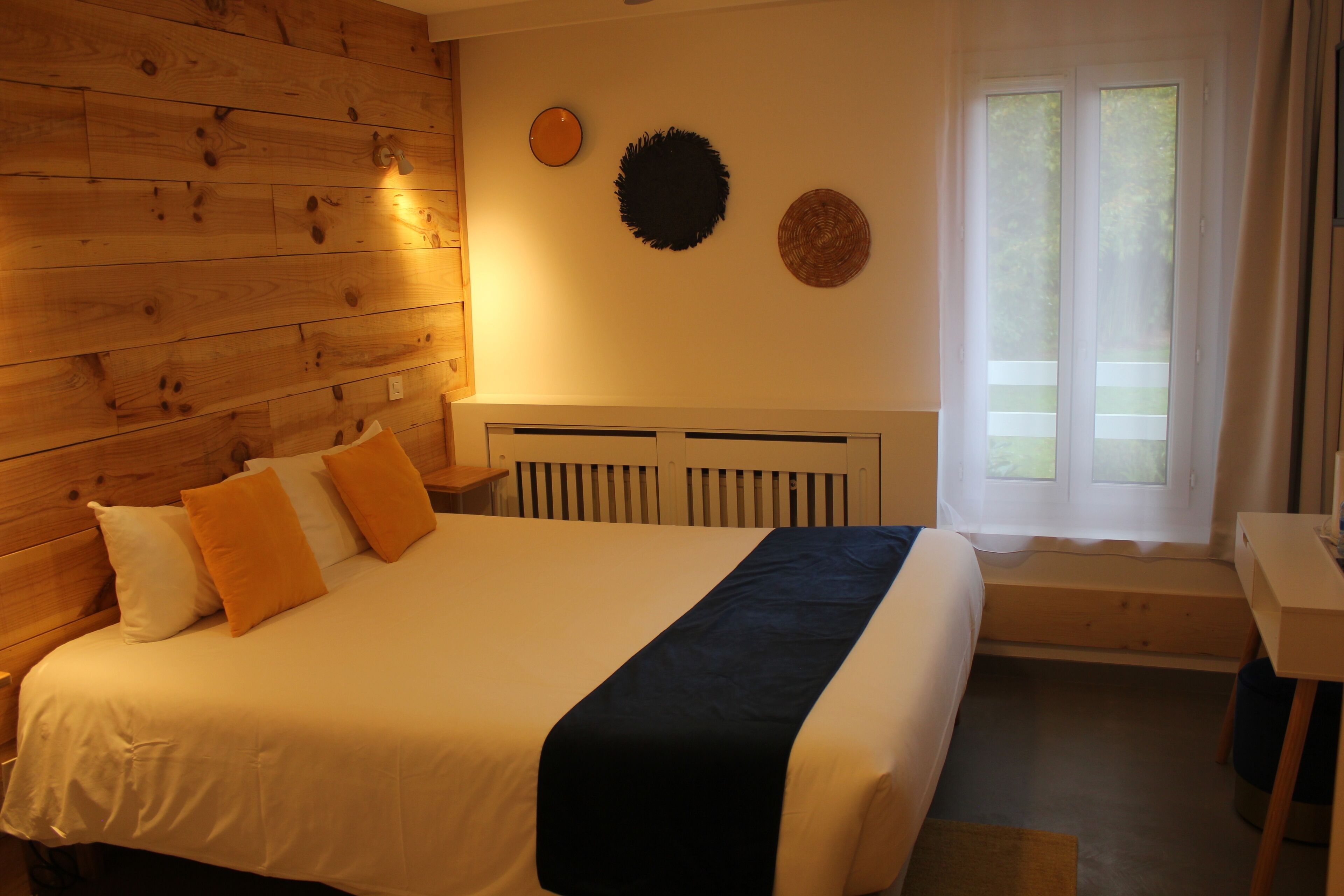 A bedroom with a wooden headboard, a bed with white and orange bedding, a black throw, and a window with a view of greenery.