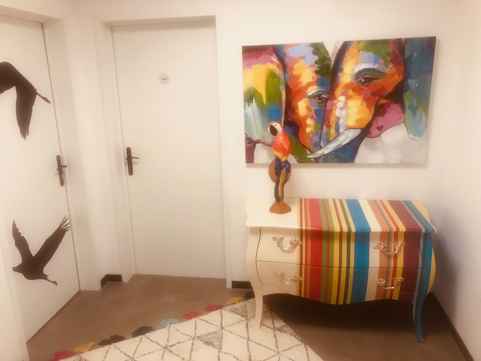 Een gang met een witte deur, een kleurrijk schilderij van een papegaai en een gestreepte tafel met lades.