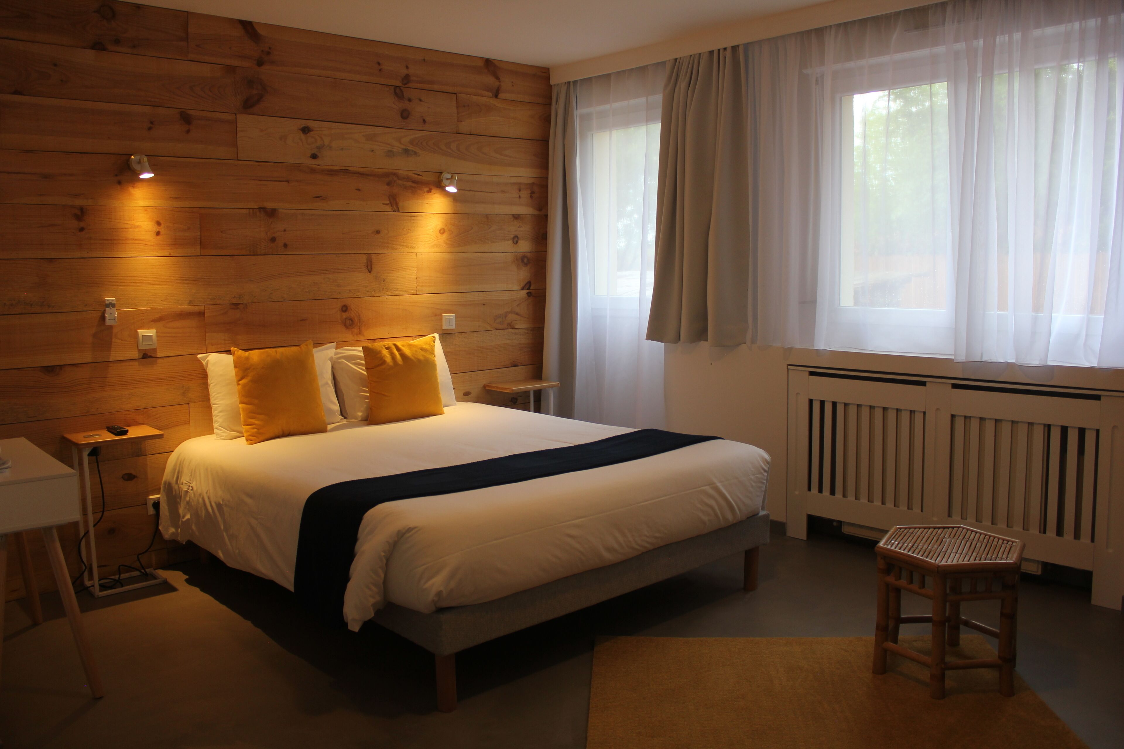 A bedroom with a wooden headboard, a bed with white and yellow bedding, a bedside table, a chair, and a window with sheer curtains.