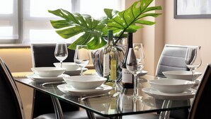 Mesa de comedor servida con platos, tazones y copas de vino blancos, junto con una botella de vino Shiraz y una planta verde grande.