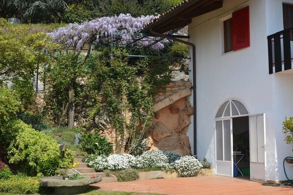 A building with a white exterior, a red shutter, and a white door. There is a garden with various plants and a tree with purple flowers. - L'Uovo e La Faina (Busca)
