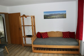 Un lit en bois avec un matelas gris et des oreillers rouges, une étagère en bois et un tableau représentant un paysage accroché au mur.