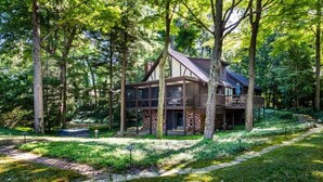 A cabin with a stone foundation, large windows, and a wooden deck, surrounded by a forested area. - HARBERT PRIVATE LAKEFRONT HOME WITH PRIVATE HOT TUB! (Harbert)