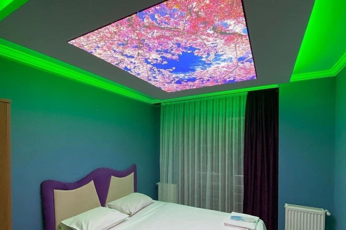 A bedroom with a bed, a window with sheer curtains, a radiator, and a ceiling with a colorful light display.