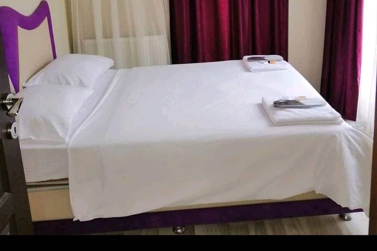 A single bed with white bedding, a purple headboard, and a small table with a folded towel and a book.