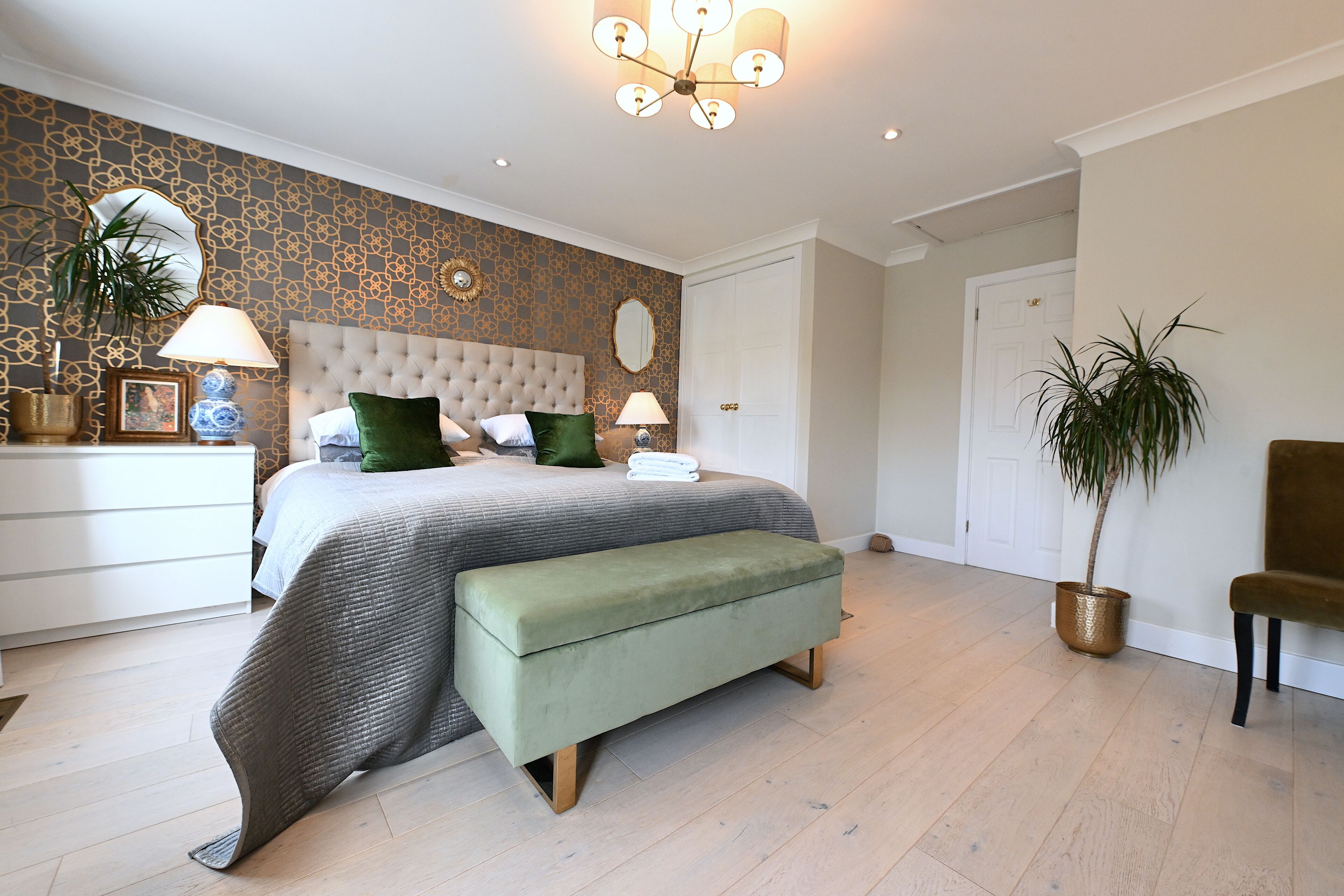 A bedroom with a tufted headboard, a large bed with green and white bedding, a bench, a potted plant, and a chandelier.