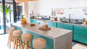 Une cuisine moderne dotée d’un îlot central, de tabourets en bois, de luminaires suspendus en fibres tressées et offrant une vue sur l’extérieur.