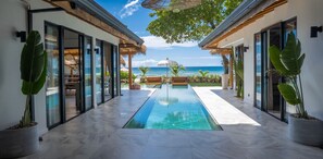 Uma área de piscina moderna com vista desimpedida do oceano, grandes vasos de plantas e uma estrutura com telhado de palha.