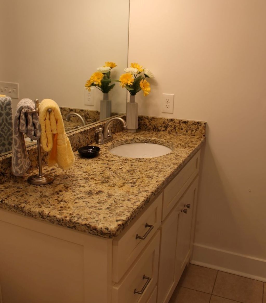 A bathroom with a granite countertop, a white sink, a towel rack with a towel, a vase with flowers, and a mirror.
