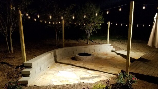 Un espace extérieur aménagé en terrasse, éclairé la nuit, avec un foyer en pierre, des marches menant à celui-ci et des guirlandes lumineuses suspendues au-dessus.