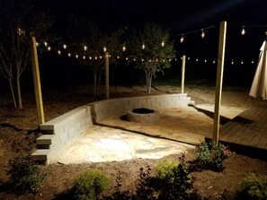 Un patio esterno serale con un braciere in pietra, gradini che vi conducono e luci a catena appese sopra.
