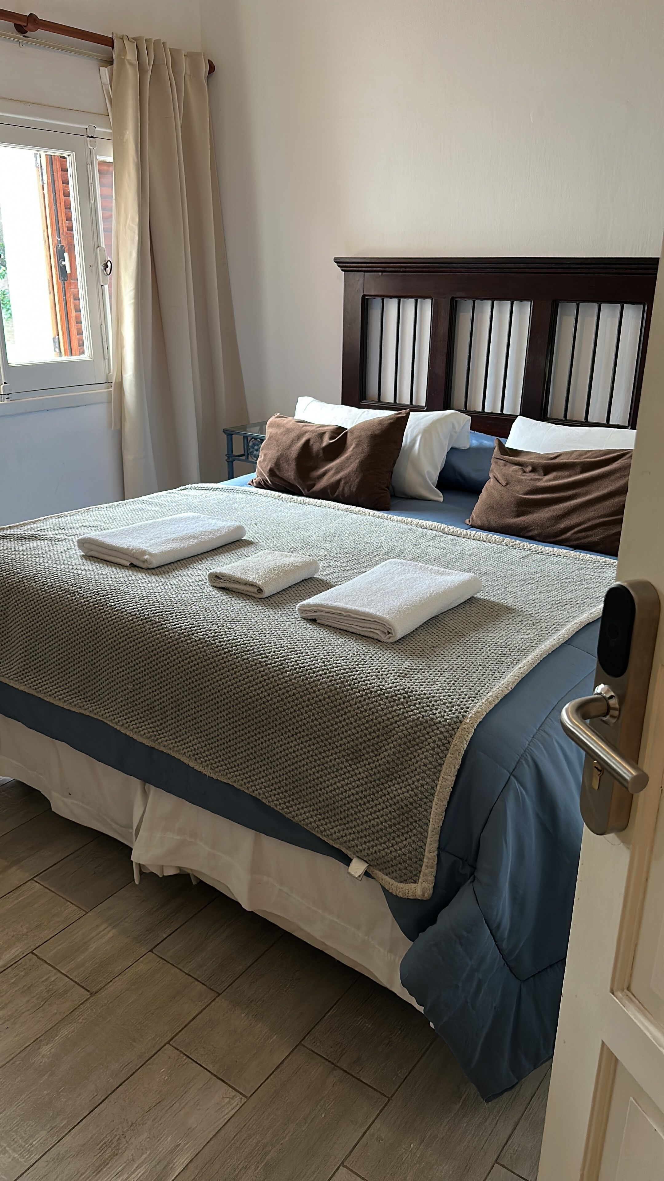 A neatly made bed with a blue and white duvet, a brown headboard, and a window with curtains.