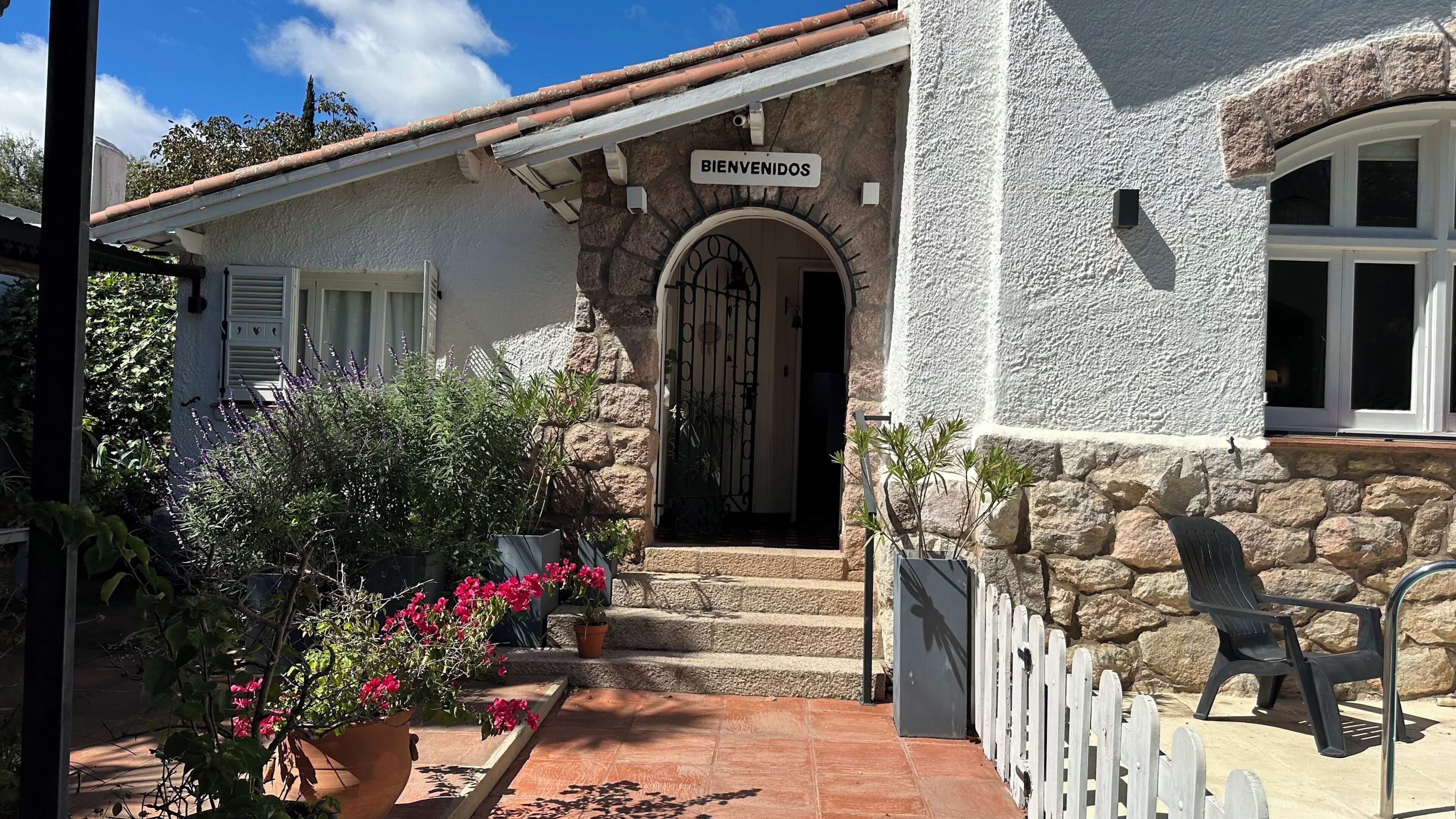 A stone building with a 'BIENVENIDOS' sign, a white picket fence, and a potted plant with pink flowers.