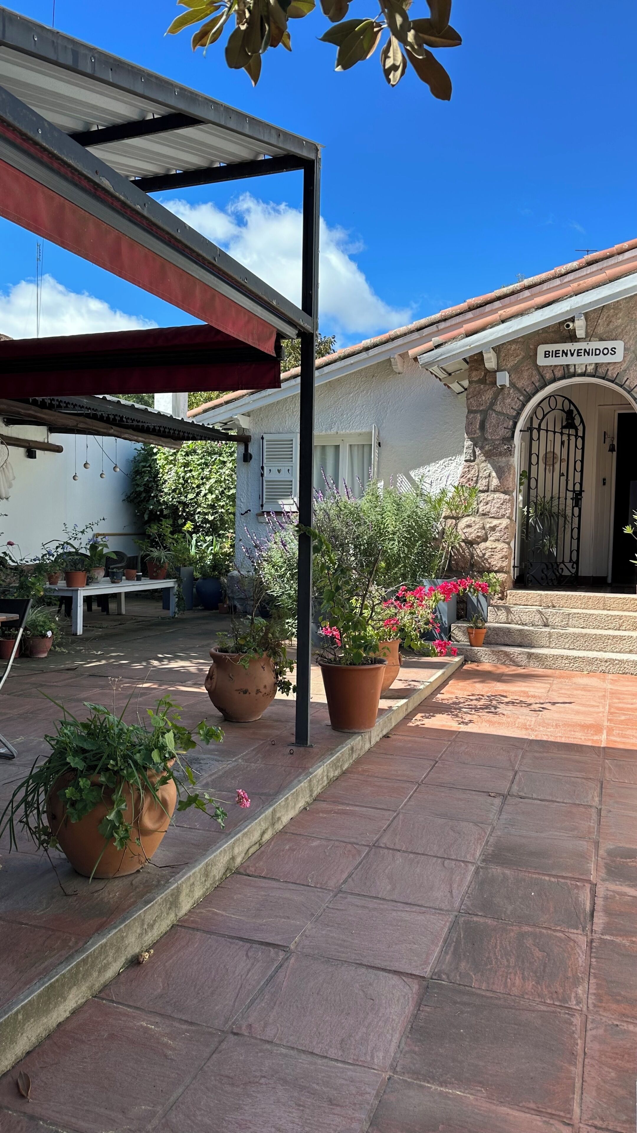 Un patio con plantas en macetas, un toldo rojo y un edificio de piedra con un cartel que dice 'BIENVENIDOS'.