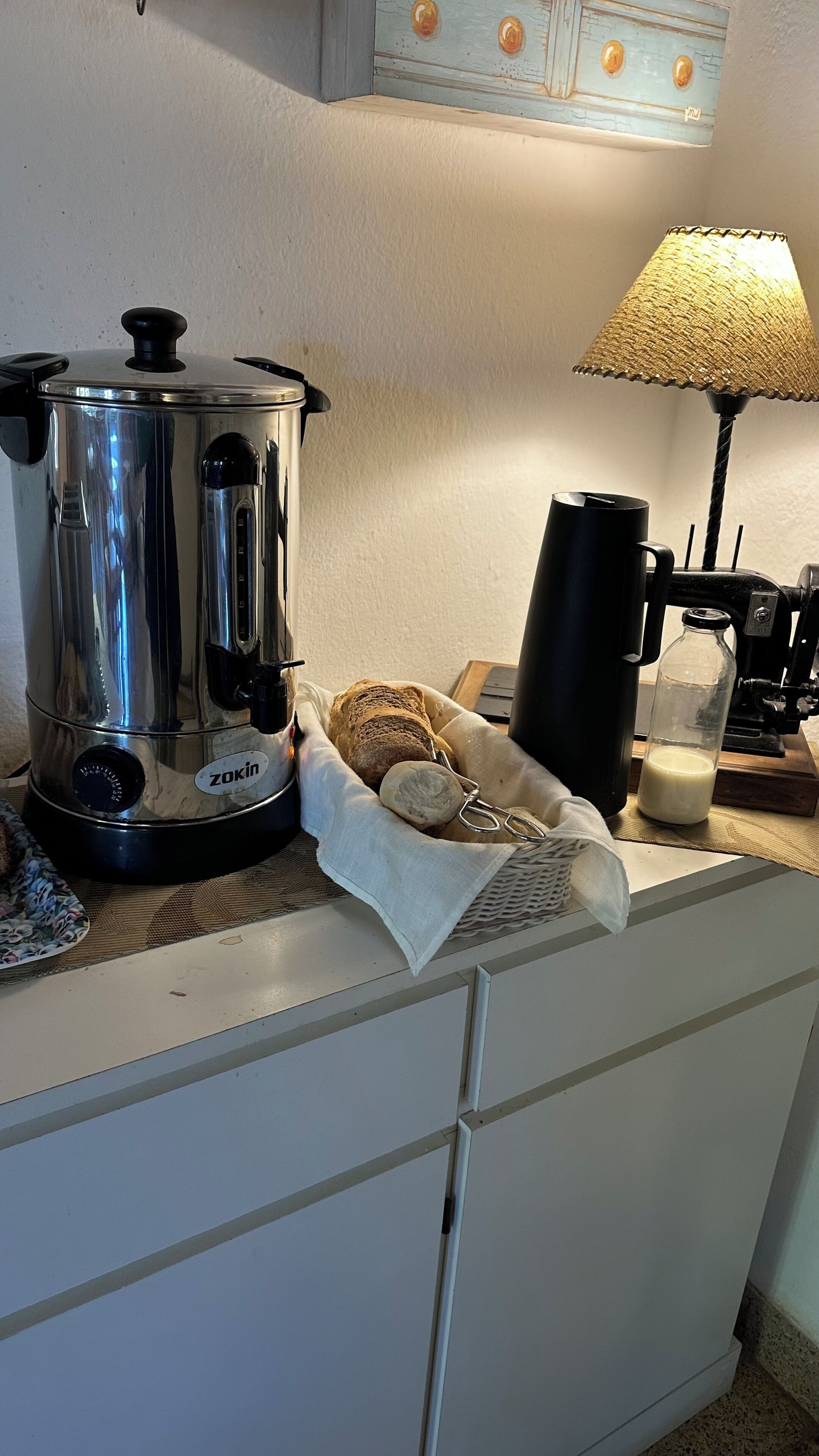 A kitchen with a stainless steel Zokin coffee maker, a black coffee pot, a bottle of milk, a loaf of bread, and a woven lamp.