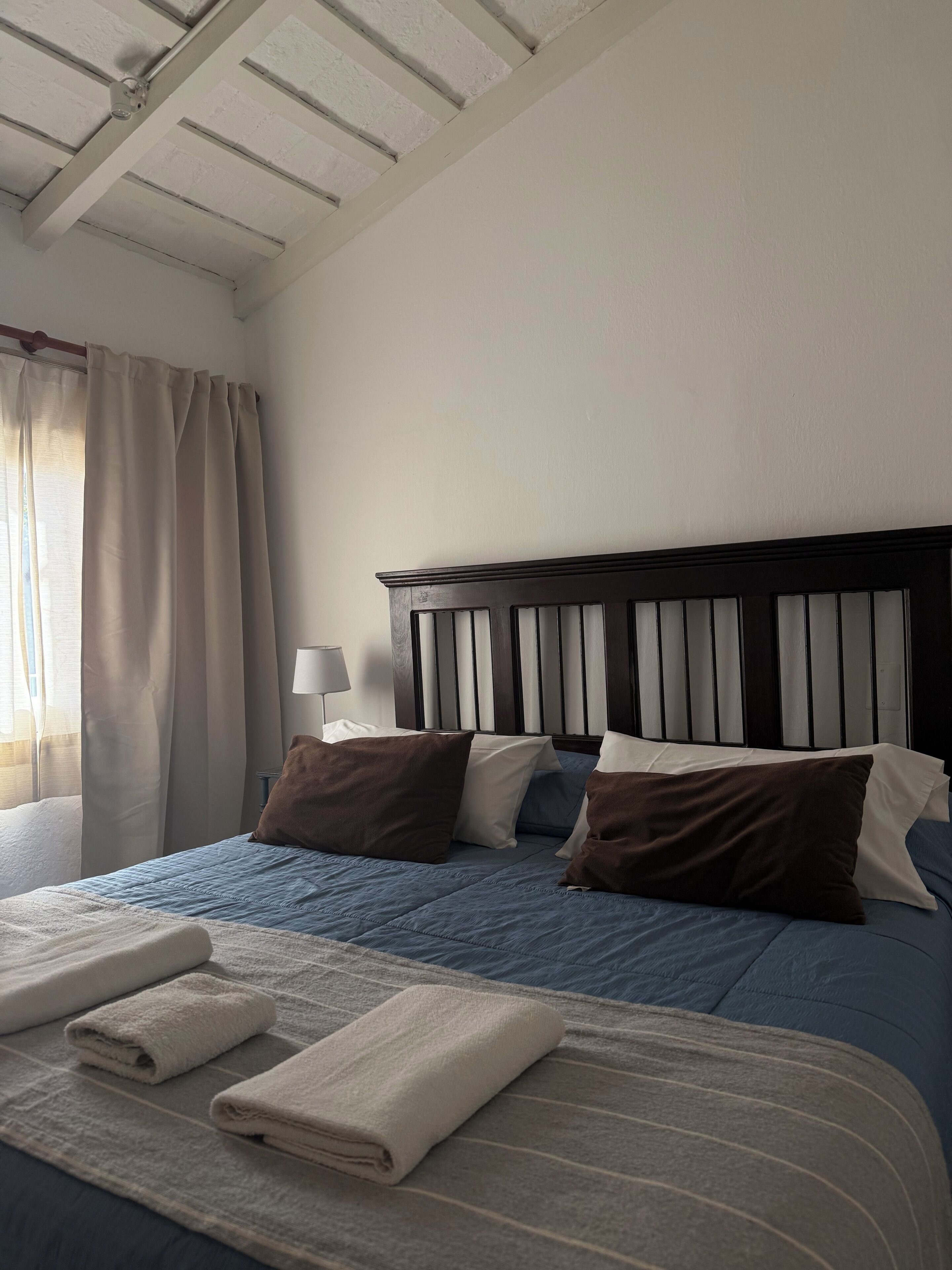 Una cama bien hecha con almohadas y una toalla doblada, una lámpara de noche y una ventana con cortinas.