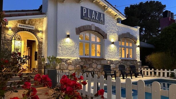 Un edificio con un cartel que dice 'PLAZA APART & HOSTFRIA', una piscina y una cerca blanca de piquetes.