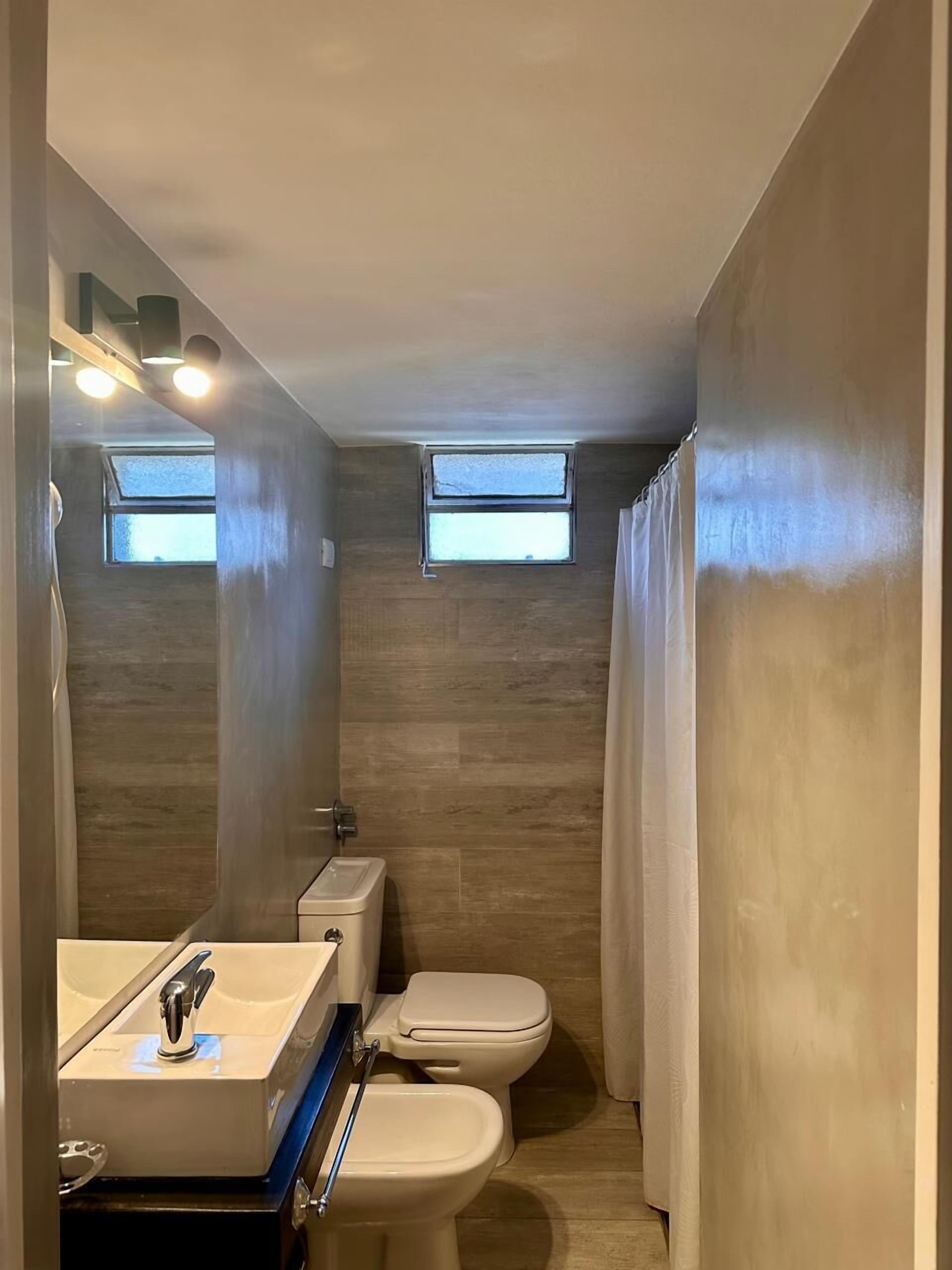 Une petite salle de bain comprenant des toilettes, un bidet et un lavabo. Elle est dotée de deux petites fenêtres et d’un rideau de douche.