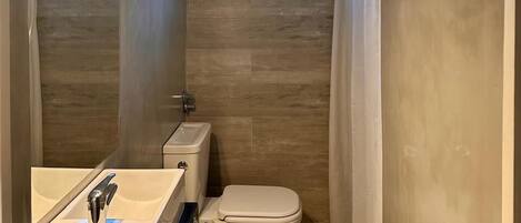 Une petite salle de bain comprenant des toilettes, un bidet et un lavabo. Elle est dotée de deux petites fenêtres et d’un rideau de douche.
