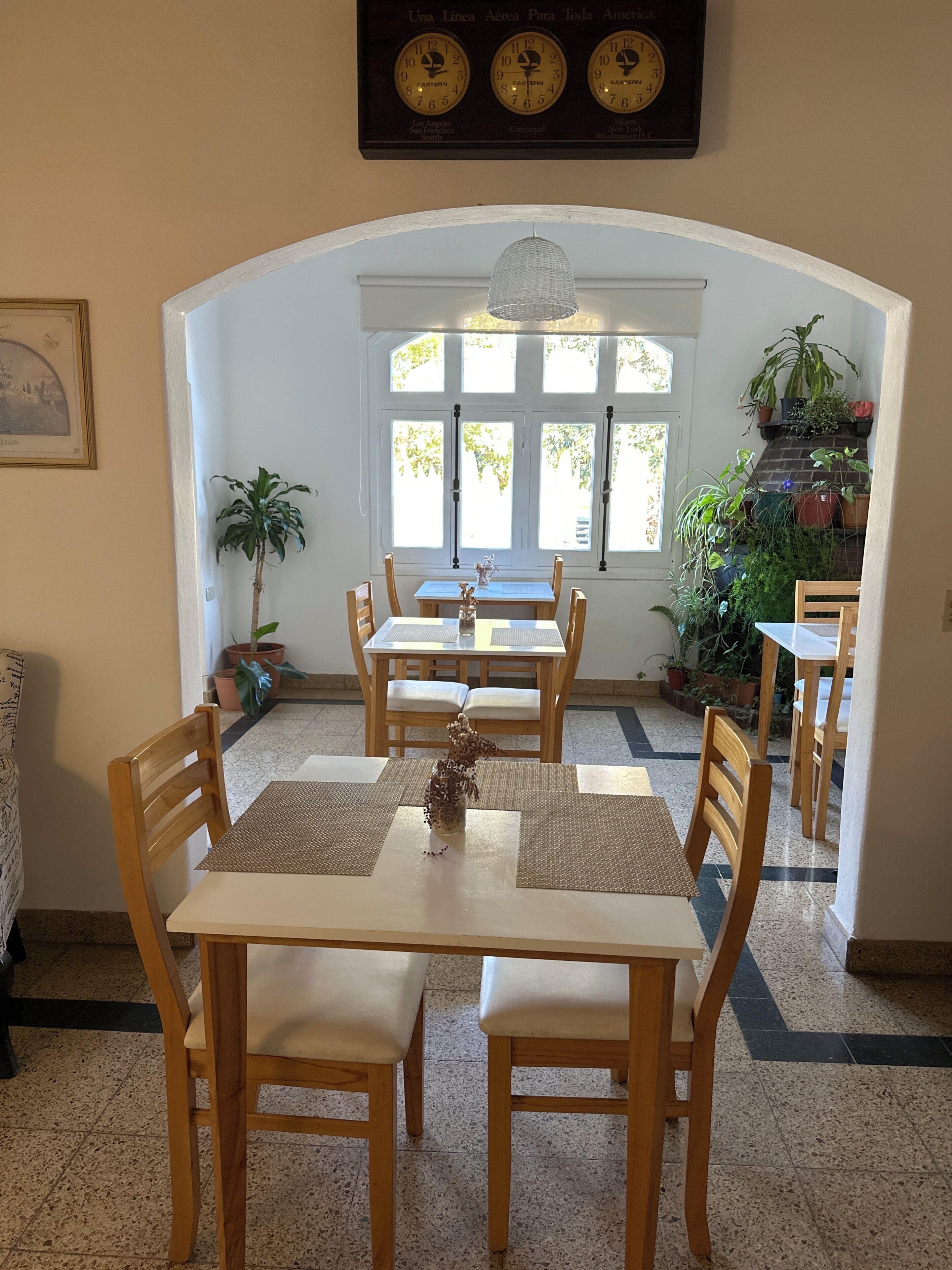 Uma área de refeições com mesas e cadeiras de madeira, um relógio acima e plantas em vasos.