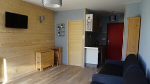 Una sala de estar con piso de madera, un sofá azul, un televisor montado en la pared y una zona de cocina con refrigerador y armarios.