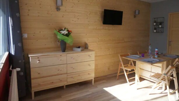 Una habitación con paneles de madera, un aparador de madera, una mesa con sillas, un televisor de pantalla plana en la pared y una ventana con cortinas.