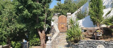 A stone pathway leading to a wooden door, surrounded by lush greenery and a stone wall.