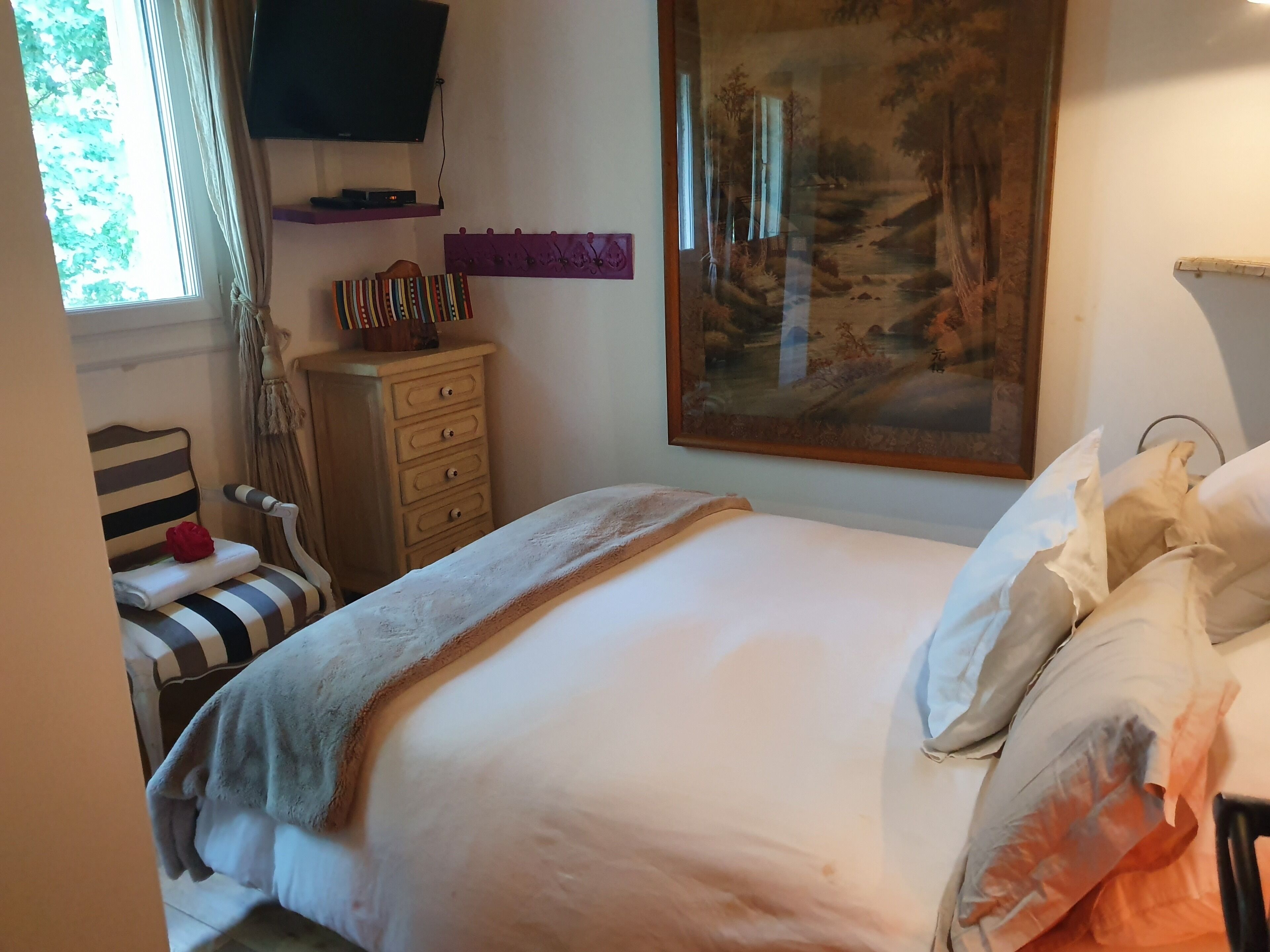 A neatly made bed with a white comforter and pillows, a wooden dresser with drawers, a striped chair, a mounted television, and a large framed painting on the wall.