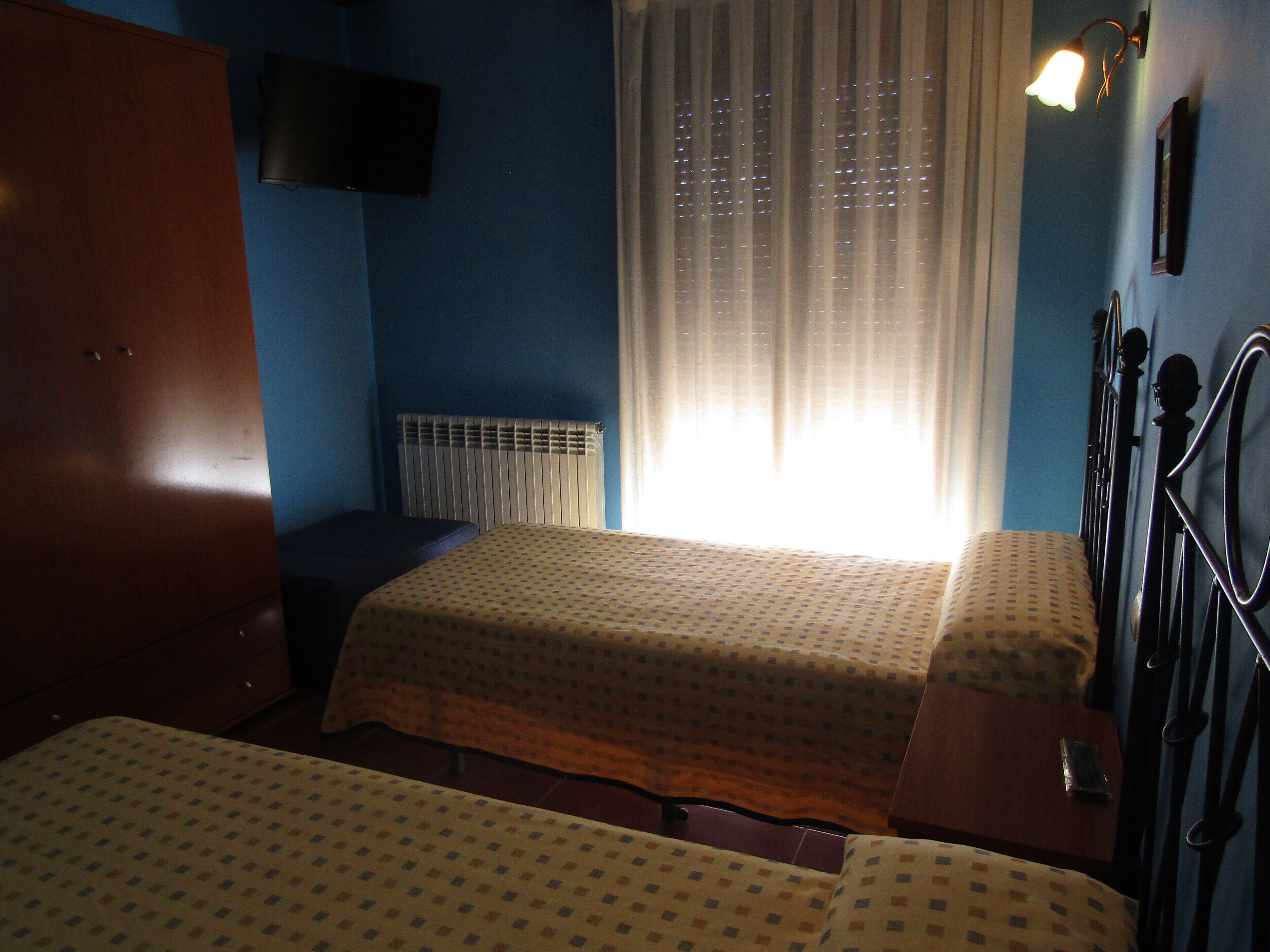 A bedroom with two beds, a wooden wardrobe, a radiator, a television, and a window with curtains.