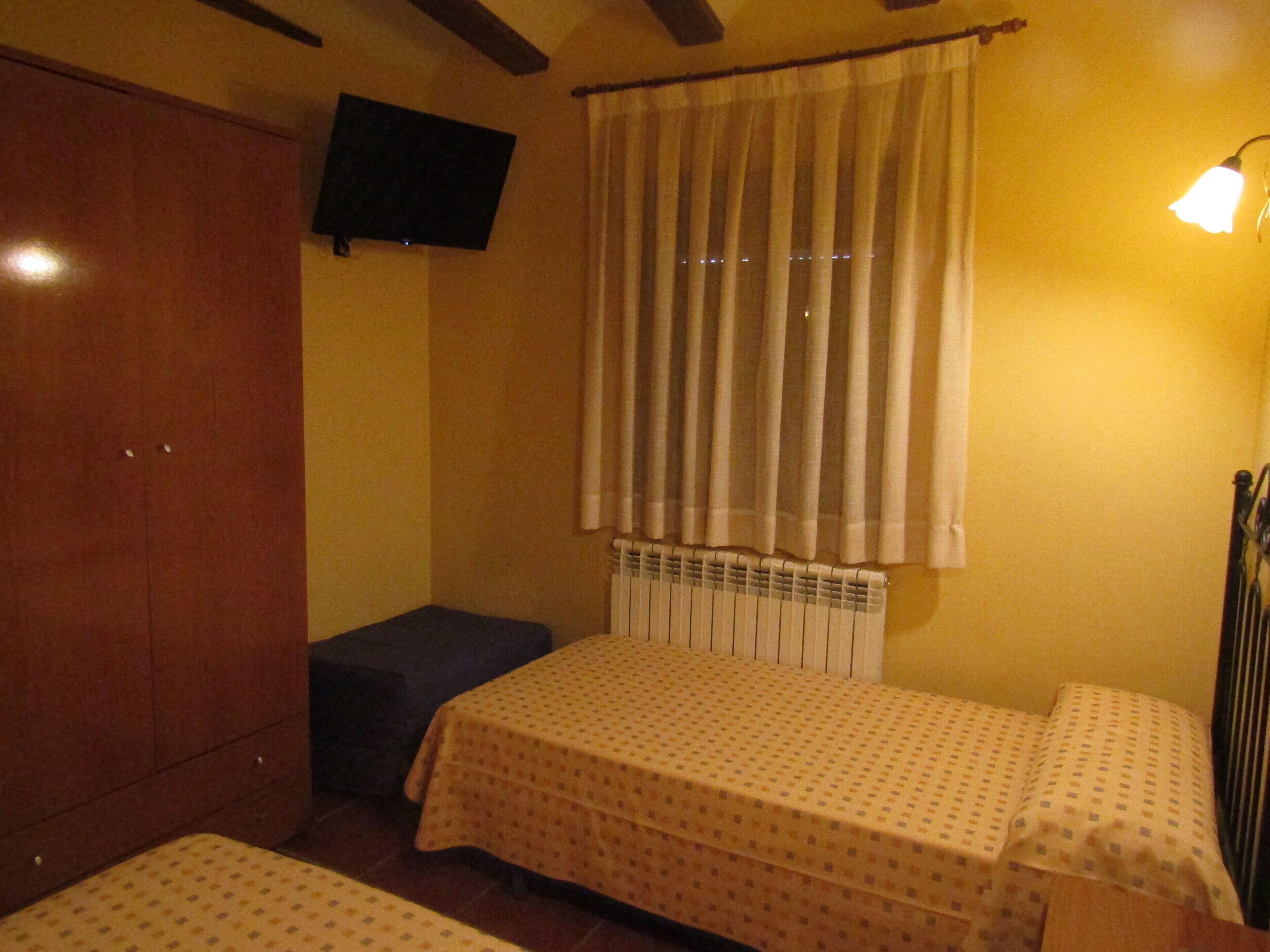 Una habitación con cama, un banco, un armario, un televisor de pantalla plana y una ventana con cortinas.