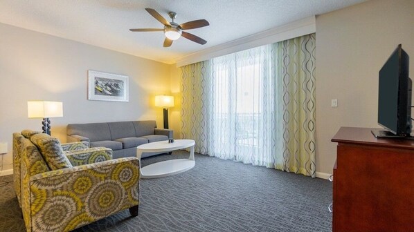 A hotel room with a sofa, armchair, TV, ceiling fan, and large window with curtains. - Life is a BEACH enjoy the WAVES! (Daytona Beach)
