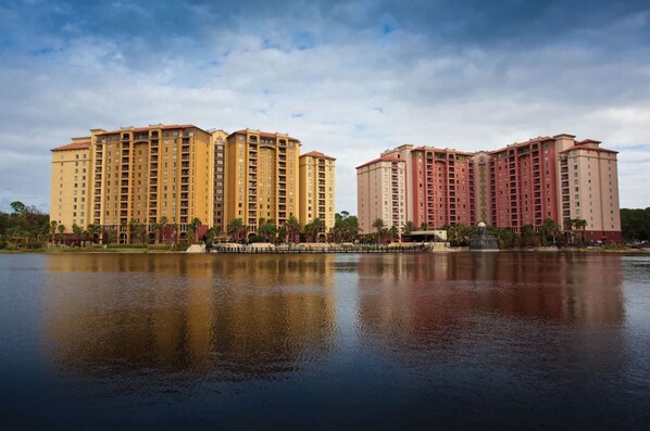 A large hotel complex with multiple high-rise buildings, a body of water in the foreground, and a clear sky. - 3BD/Orlando Near Disney - 5 Pools, Game Room, Café and Restaurant On-Site! (Lake Buena Vista)