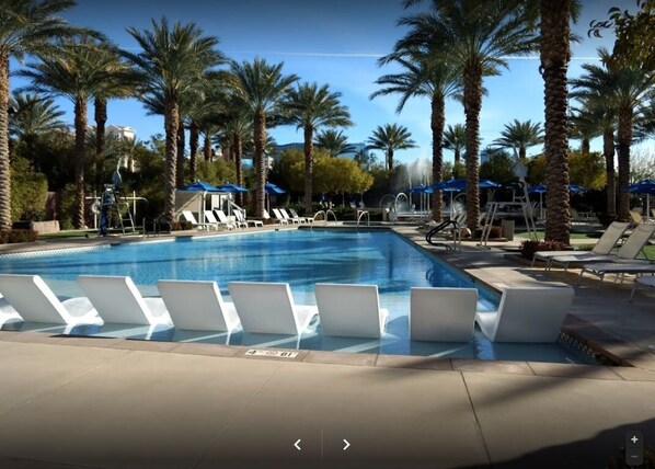 Relax by the pool surrounded by palm trees and lounge chairs, with umbrellas and a soothing water fountain nearby.
