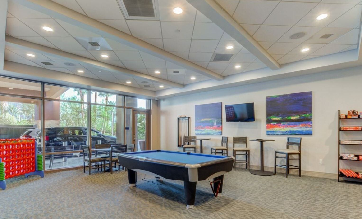 Une table de billard dans une pièce bien éclairée, dotée de grandes fenêtres, d’un téléviseur fixé au mur et d’une bibliothèque.