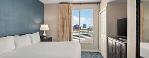 A hotel room with a bed, a television, a nightstand, a lamp, a window with curtains, and a view of a cityscape.
