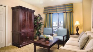 Sala de estar con un mueble de madera oscura, dos sillones, un sofá, una mesa de centro con flores, una lámpara y vistas a un paisaje urbano a través de amplias ventanas con cortinas a rayas.