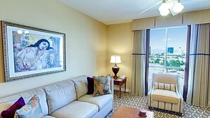 A hotel room with a sofa, a chair, a table, a lamp, a framed painting, and a view of the city through large windows.