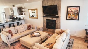 A modern living room with a sofa set, a fireplace, a television, and a kitchen in the background. - Downtown BV Retreat _311_ Steps to Main & River (Buena Vista)