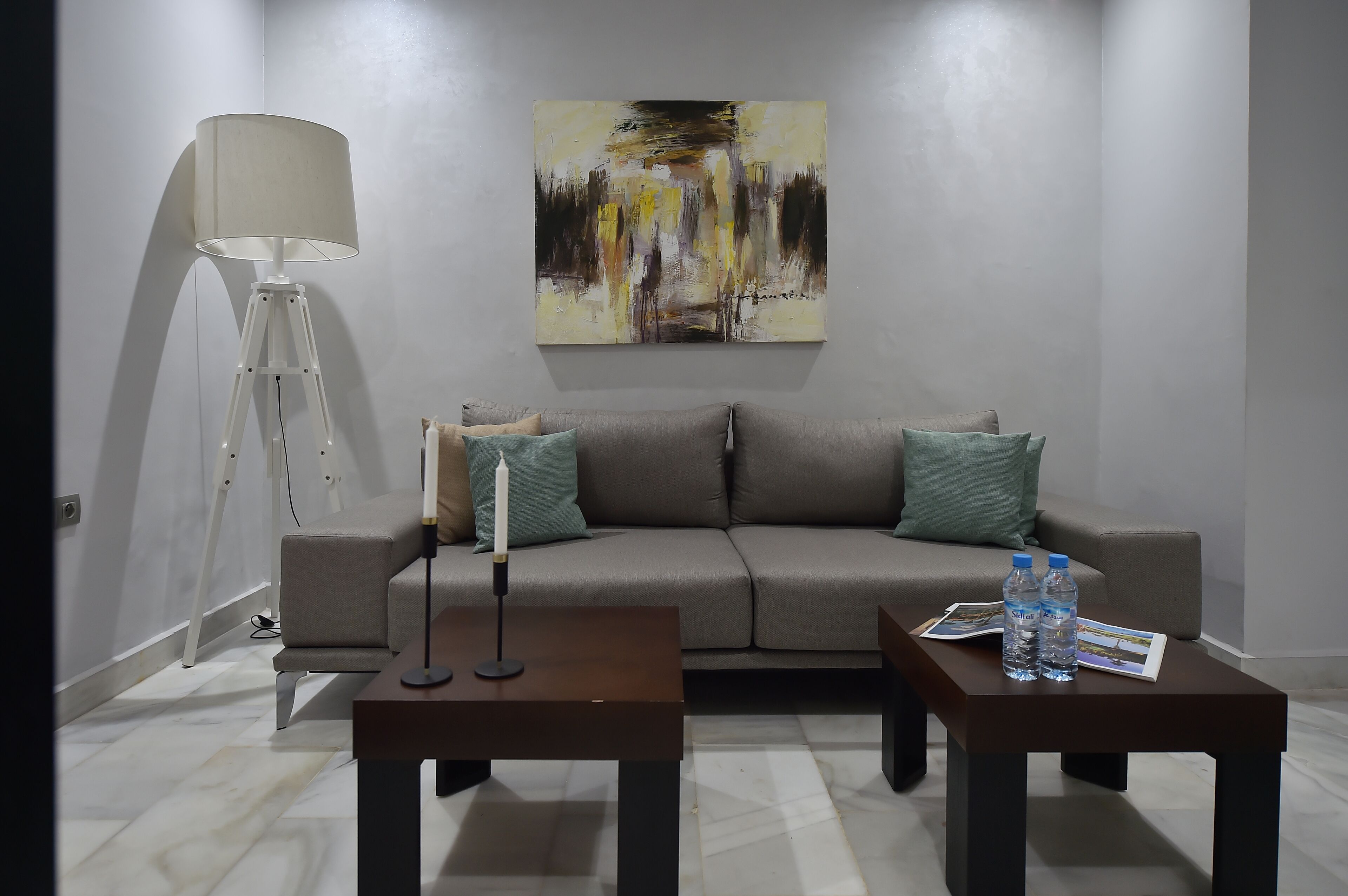 Ein modernes Wohnzimmer mit einem grauen Sofa, zwei Beistelltischen, einer Stehlampe und einem großen, abstrakten Gemälde an der Wand.