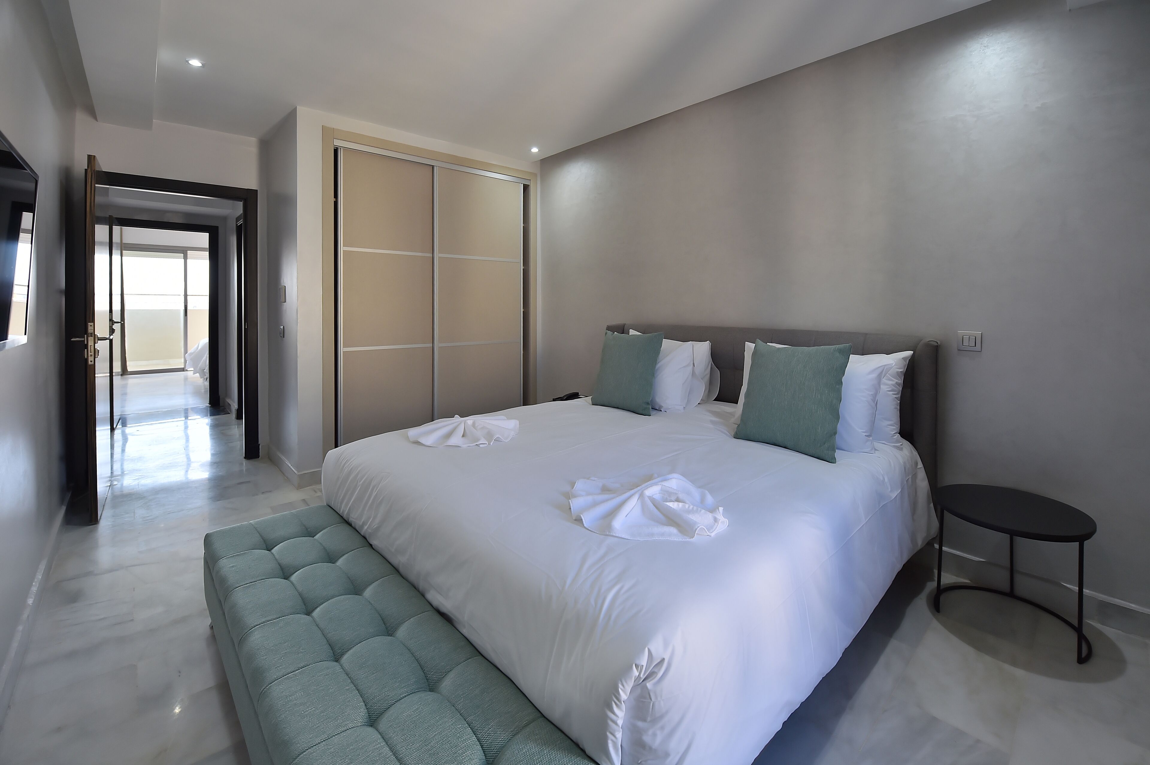 A modern hotel room with a large bed, a bench, a wardrobe, and a small table.