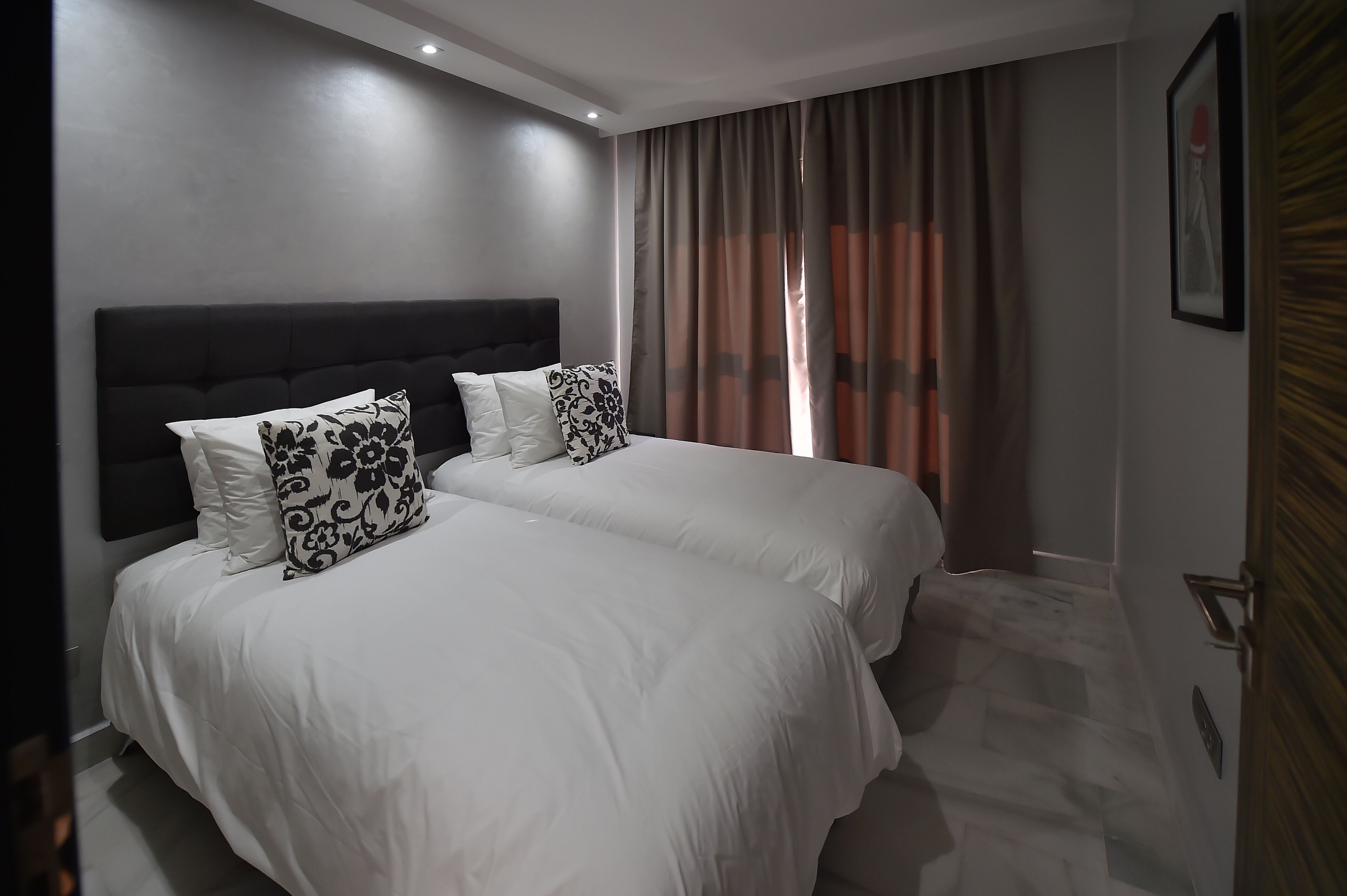 Habitación de hotel con dos camas, cabecera oscura y una ventana grande con cortinas.