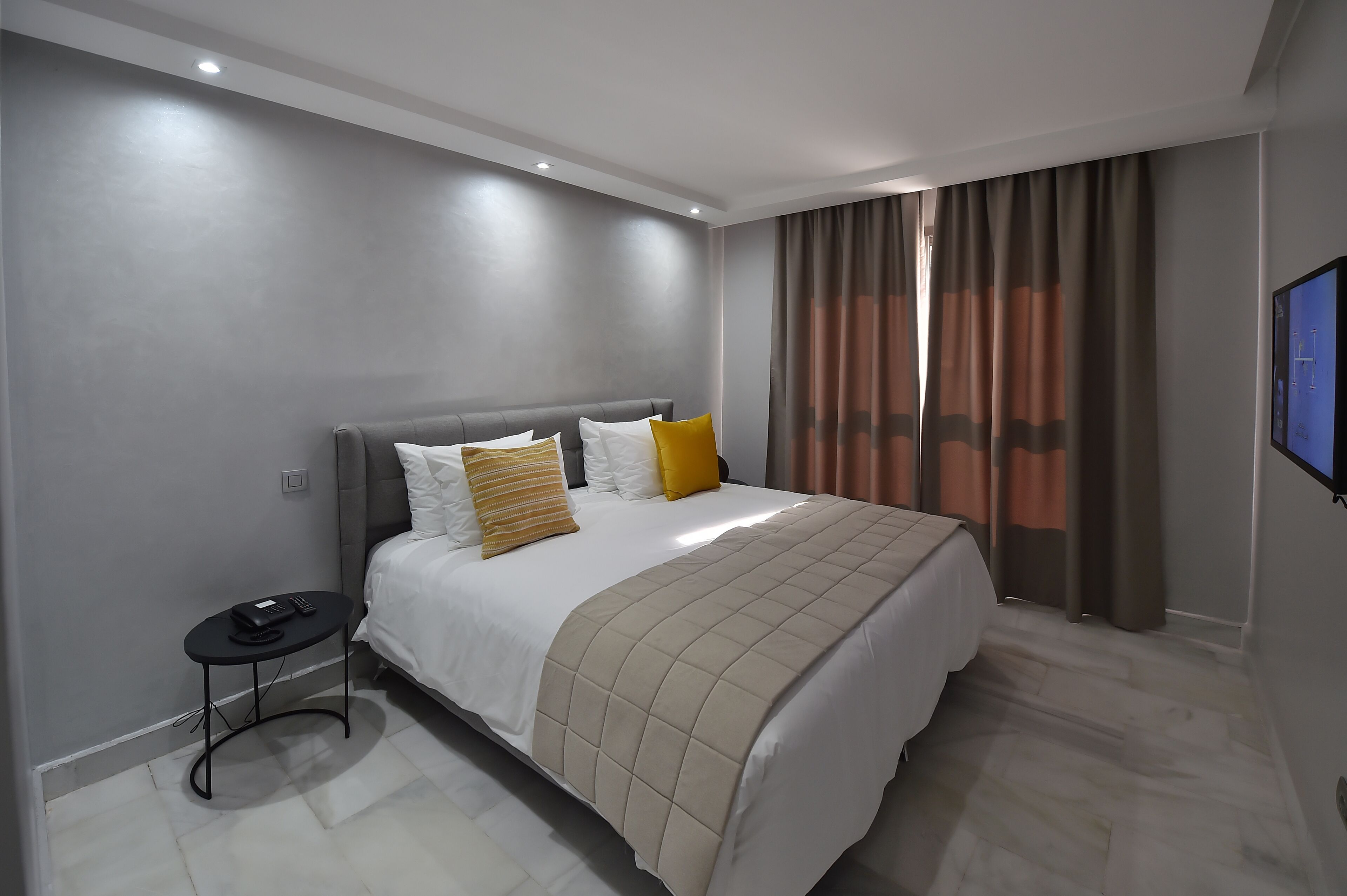 A modern hotel room with a large bed, a bedside table with a phone, and a TV on the wall.